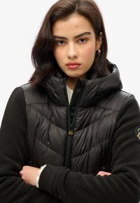 Chaqueta acolchada negra con un diseño de colchas, cuello alto y cierre frontal con cremallera. Presenta mangas de forro polar suave y detalles de hardware en tono dorado.