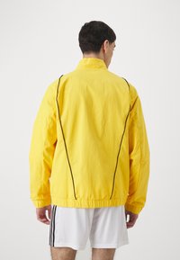 Chaqueta ligera amarilla con costuras negras, cuello alto y puños elásticos. Textura suave con un corte holgado, vista desde la parte posterior.