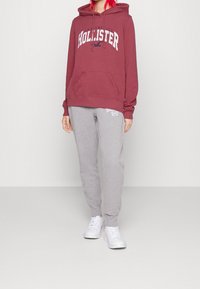 Sudadera color marrón con el logo de "Hollister", bolsillo tipo canguro y capucha con cordón; combinada con pantalones de chándal grises y zapatillas blancas.