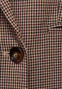 Vlnená sako látka s vzorom houndstooth v odtieňoch burgundskej, smotanovej a čiernej; obsahuje hnedý gombík a úzke klopy.