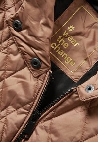 Brun quiltet jakke med sort lynlås, sorte trykknapper og en gylden label indeni, der står "#wear the change."
