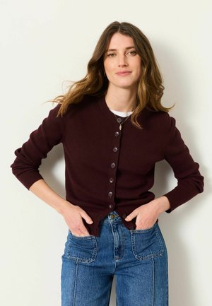 Woman with long brown hair wearing a dark buttoned cardigan and blue jeans with large front pockets, standing with hands in pockets.
