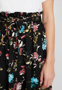 Zwarte bloemenrok met gerimpelde tailleband, voorzien van kleurrijke bloemen in roze, blauw en goud. Strikdetail op de taille.