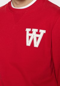 Červená mikina vyrobená z bavlny s kulatým výstřihem, která má výrazné bílé vyšívané logo "W" na levé části hrudi.