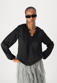 Blusa nera a maniche lunghe con scollatura a V, caratterizzata da dettagli in pizzo intricati. Tessuto morbido, vestibilità rilassata e maniche leggermente a sbuffo.