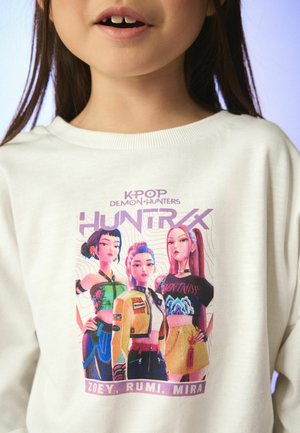 Tüdruk kannab valget pusa, millel on kujutatud kolm animeeritud K-popi tegelast värvikate riietustega ning allpool nimed Zoey, Rumi, Mira.