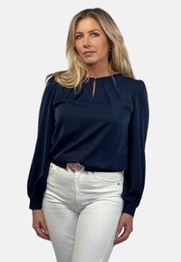 Granatowa bluzka z długim rękawem z plisą przy szyi, noszona z białymi dżinsami i czarnym paskiem z srebrną klamrą.