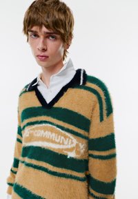 Fuzzový sveter s výstrihom do V v béžovej farbe so zelenými a námorníckymi pruhmi, s bielym logom v strede. Mäkkučký, textúrovaný materiál s voľným strihom.