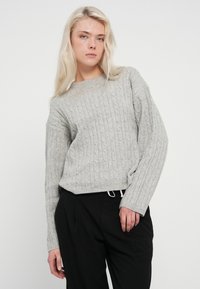 Világosszürke kötött pulóver bordázott mintázattal, kerek nyakkivágással és lezuhant vállakkal, magas derekú fekete nadrággal párosítva.