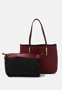 Burgundská taška přes rameno s odnímatelnou černou kapsou, která má bordó lem a zipové uzavírání, obě stojící na bílém pozadí.
