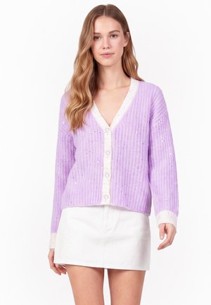 Ung kvinde med langt, bølget hår iført en lavendelfarvet cardigan med knapper og hvide kanter samt en hvid mininederdel mod en ensfarvet baggrund.