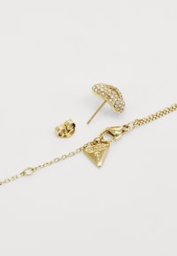 Kuldseteid ja kaelakeed, millel on särav ja tekstuuritatud disain koos rhinestone'idega ning kolmnurkne charm brändinimega.
