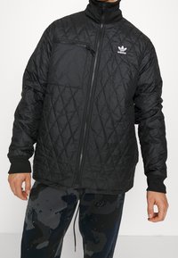 Svart kviltad jacka med ståkrage, utrustad med en dragkedjeförsedd bröstficka och stickade ärmslut. Logotyp på vänster bröst. Lättviktsmaterial.