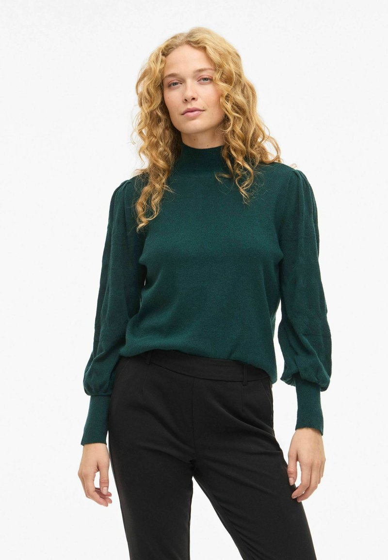 Maglione verde a maniche lunghe con collo alto, tessuto a trama, e maniche a sbuffo, abbinato a pantaloni neri su misura.