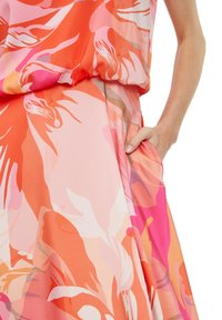 Kvinnas hand vilar i fickan på en klänning med orange, rosa och vit blommig mönstring och en detaljerad midja med rynkor.