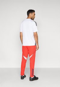 Biały sportowy t-shirt z czarnymi bocznymi paskami, w zestawie z jaskrawoczerwonymi spodniami z odblaskowymi akcentami i czarnymi trampkami.