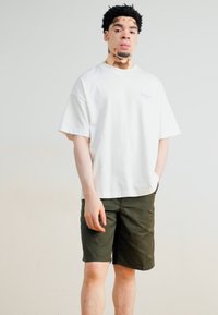 Tricou alb din bumbac, cu decolteu rotund și mâneci scurte, asortat cu pantaloni scurți de culoare verde măsliniu. Tricoul are detalii brodate subtile.