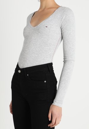 Mujer vistiendo una camiseta ajustada de manga larga color gris claro con un pequeño logotipo y pantalones negros de cintura alta, sobre un fondo liso.