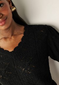 Top nero lavorato a maglia con scollo ondulato, texture a motivo e maniche a sbuffo. Presenta disegni a forma di diamante su tutta la superficie.