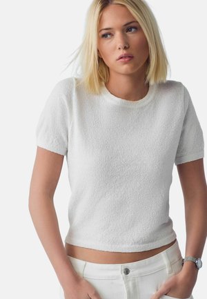 Młoda kobieta o blond włosach, ubrana w biały krótki teksturowany top i białe spodnie, lekko patrzy na bok, mając ręce w kieszeniach.