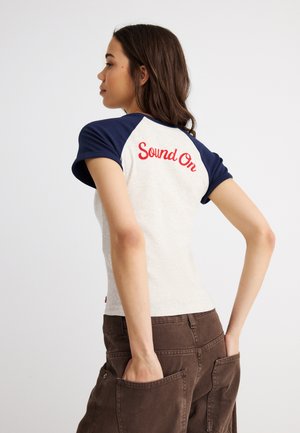 Junge Frau, die nach links schaut, trägt ein weißes und marineblaues Hemd mit rotem "Sound On"-Text auf dem Rücken, die Hände in den großen braunen Hosentaschen.