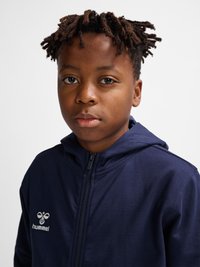 Dreng med kort, snoet hår iført en mørkeblå lynlåshættetrøje med et hummel-logo, der kigger fremad med et neutralt udtryk.