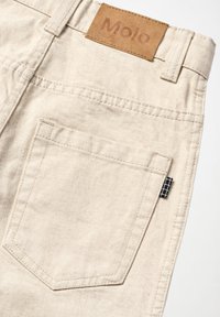 Pantalones de mezclilla beige con bolsillo trasero, trabillas para el cinturón, etiqueta a cuadros en blanco y negro y parche de cuero marrón etiquetado "Molo" en la cintura.