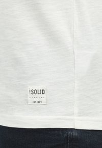 Fehér, puha anyagból készült póló, alján egy márkás címkével, amelyen a "ISOLID DENMARK EST.1999" felirat szerepel fekete szöveggel.