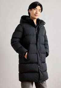 Schwarze Pufferjacke mit Kapuze, ausgestattet mit einem gesteppten Design, Vorderknöpfen und seitlichen Taschen. Der Stoff wirkt glatt mit einem matten Finish.