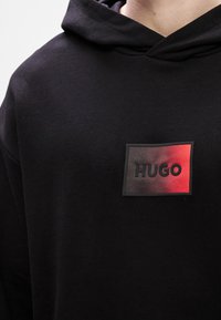 Μαύρη κουκούλα με ορθογώνια ετικέτα που �γράφει "HUGO" μαύρα σε φόντο με κλίση κόκκινο. Μαλακό υλικό και άνετος σχεδιασμός.