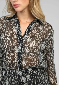 Blouse transparente avec un motif abstrait noir et blanc, dotée d'un col classique, d'une fermeture à boutons sur le devant et de manches longues. Tissu léger et doux.