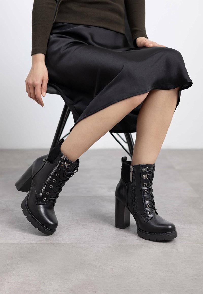 Botas de cuero negras hasta el tobillo con un tacón grueso, parte frontal con cordones, cierre de cremallera y suela texturizada. Combinadas con una falda de satén negra.