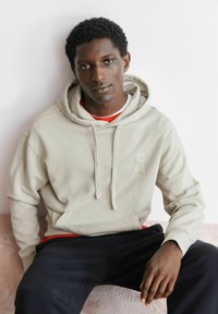 Lichtbeige hoodie met trekkoorden, voorzien van een klein logo. Gedragen over een rood shirt, gecombineerd met zwarte broek. Gladde stof en losse pasvorm.