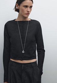 Svart, kortärmad lång cardigan med knäppning framtill, parat med höga svarta byxor. Silversmycke med hänge ger en detaljerad touch.