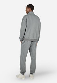 Vyras stovi su rankomis kišenėse, apsirengęs pilku sportiniu džemperiu ir derančiomis sportinėmis kelnėmis, su baltais sportbačiais ant balto fono.