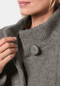 Gray wool coat with a textured finish, featuring a large gray button with four holes. Hand rests on the collar.