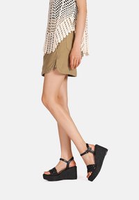 Sandali wedge neri con una piattaforma testurizzata, cinturino alla caviglia regolabile e dettagli intrecciati sulla parte superiore. Indossati con pantaloni corti color khaki e un top in pizzo.
