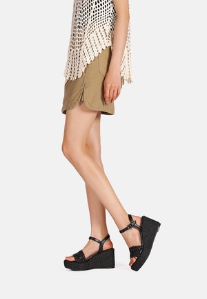 Sandali wedge neri con una piattaforma testurizzata, cinturino alla caviglia regolabile e dettagli intrecciati sulla parte superiore. Indossati con pantaloni corti color khaki e un top in pizzo.