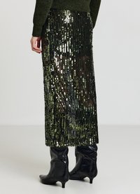 Jupe noire à paillettes coupe droite, ornée de paillettes vertes et argentées scintillantes formant des lignes verticales, accompagnée de bottes noires montant jusqu'aux genoux.