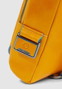 Gele leren tas met een gestructureerde afwerking, voorzien van een blauw accent bij de rits en een metalen logoplaatje bevestigd.