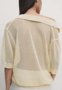 Femme de dos portant un haut en maille beige clair, ample, avec un col large glissant sur une épaule, laissant apparaître la peau en dessous.