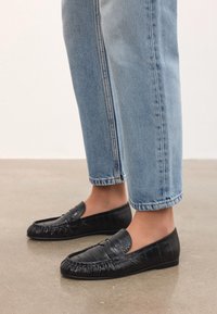 Mustad nahast loafers tekstureeritud äärisega, lame talla ja sileda pealmise osaga. Kombineeritud helesiniste lühikeste teksadega.