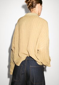 Beige texturovaná košile s volným střihem a límečkem, v kombinaci s tmavými džíny s kapsami a lehce elastickým pasem.