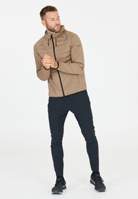 Leichte, tanfarbene Jacke mit taillierter Schnittführung, Reißverschluss und verstellbarer Kapuze, kombiniert mit schwarzen Sporthosen und dunklen Schuhen.