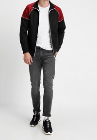 Giacca nera con zip, dettagli rossi sulle spalle e piping bianco, abbinata a jeans grigi e sneakers nere, esposta su una figura in piedi.