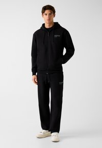 Sweat à capuche noir zippé avec une poche kangourou et une capuche ; assorti à un pantalon de jogging assorti. Les deux présentent des accents discrets du logo "GUESS".