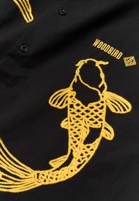 Tissu noir avec un poisson koi jaune brodé, le mot "WOODBIRD" et un petit logo géométrique près de boutons noirs.