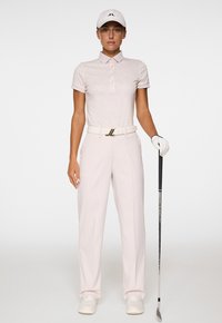 Donna in un abbigliamento da golf rosa pallido, con un berretto bianco e un guanto, tiene una mazza da golf contro uno sfondo bianco semplice.