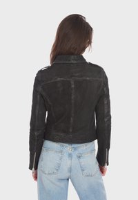 Giacca di pelle nera con design aderente, spalline e cerniere in argento. Finitura texturizzata, lunghezza corta, abbinata a jeans azzurri.