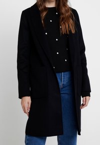 Černý dlouhý kabát se širokými klopami, dvěma předními kapsami a rovný střihem, doplněný černým svrškem s puntíky a modrými džínami.